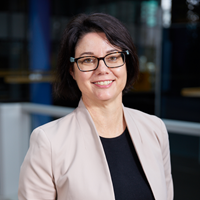 Woman with short brown hear wearing glasses, beige blazer and back top smiling at camera for profile