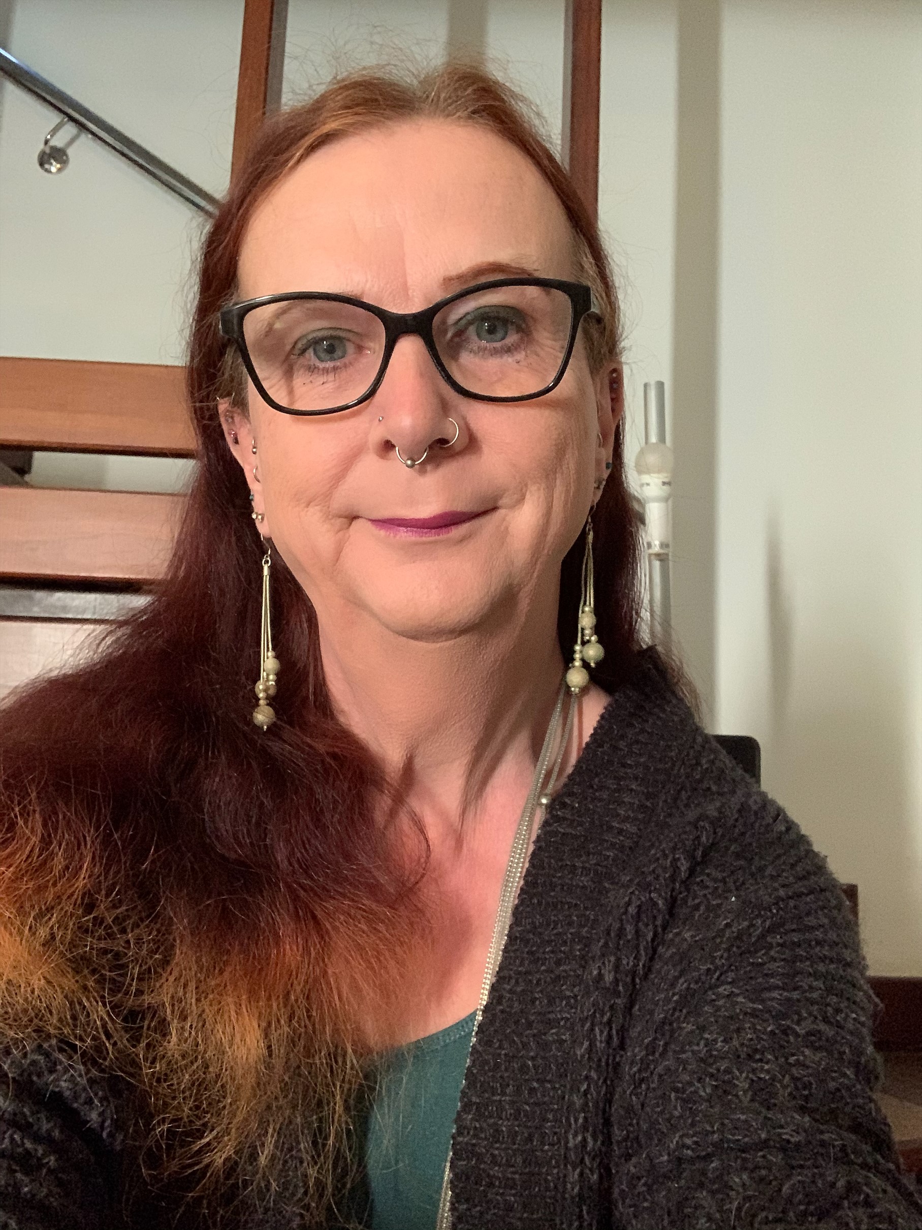 A woman wearing glasses, earrings and a dark grey jumper taking a selfie photo