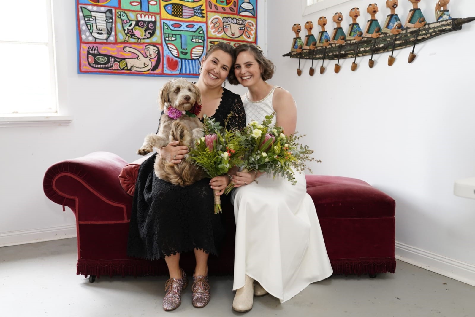 two women at their wedding sitting on a red velvet couch with their dog