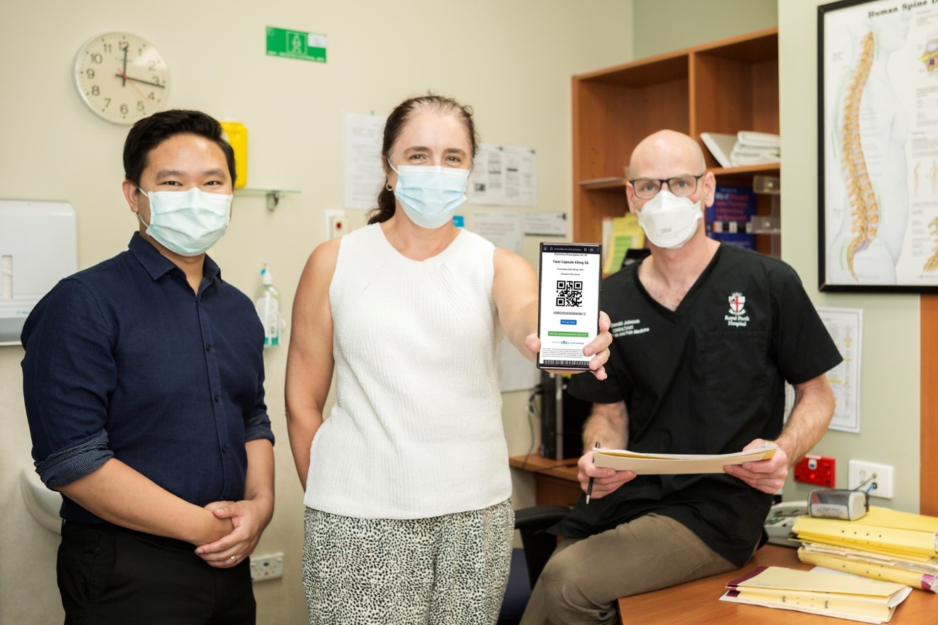 Three people with face masks on posing for a photo in a medical room at RPH. One lady holds a phone showing the RPH Electronic Prescribing app