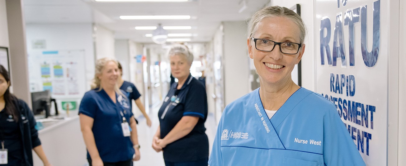 A Nurse standing in a hospital corridor smiling with staff nearby watching on